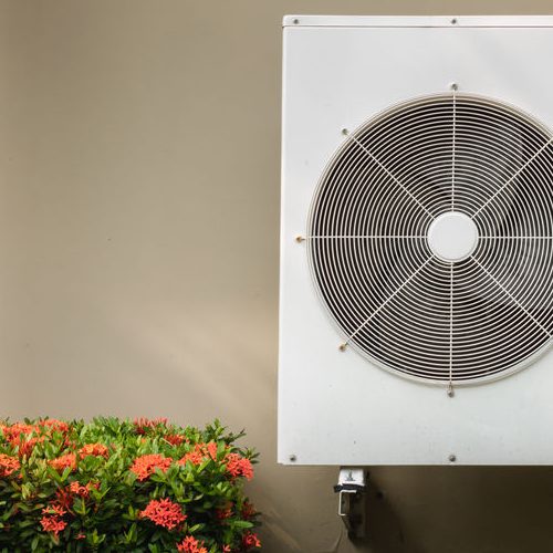 Compressor air conditioning system installation embedded on wall of building with flowers in a shrub on the left hand side of the photo.